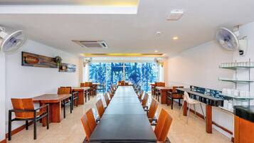Un vaste espace dédié à la salle à manger, avec de longues tables et des chaises, des ventilateurs de plafond et une vue sur les arbres à travers de grandes fenêtres.