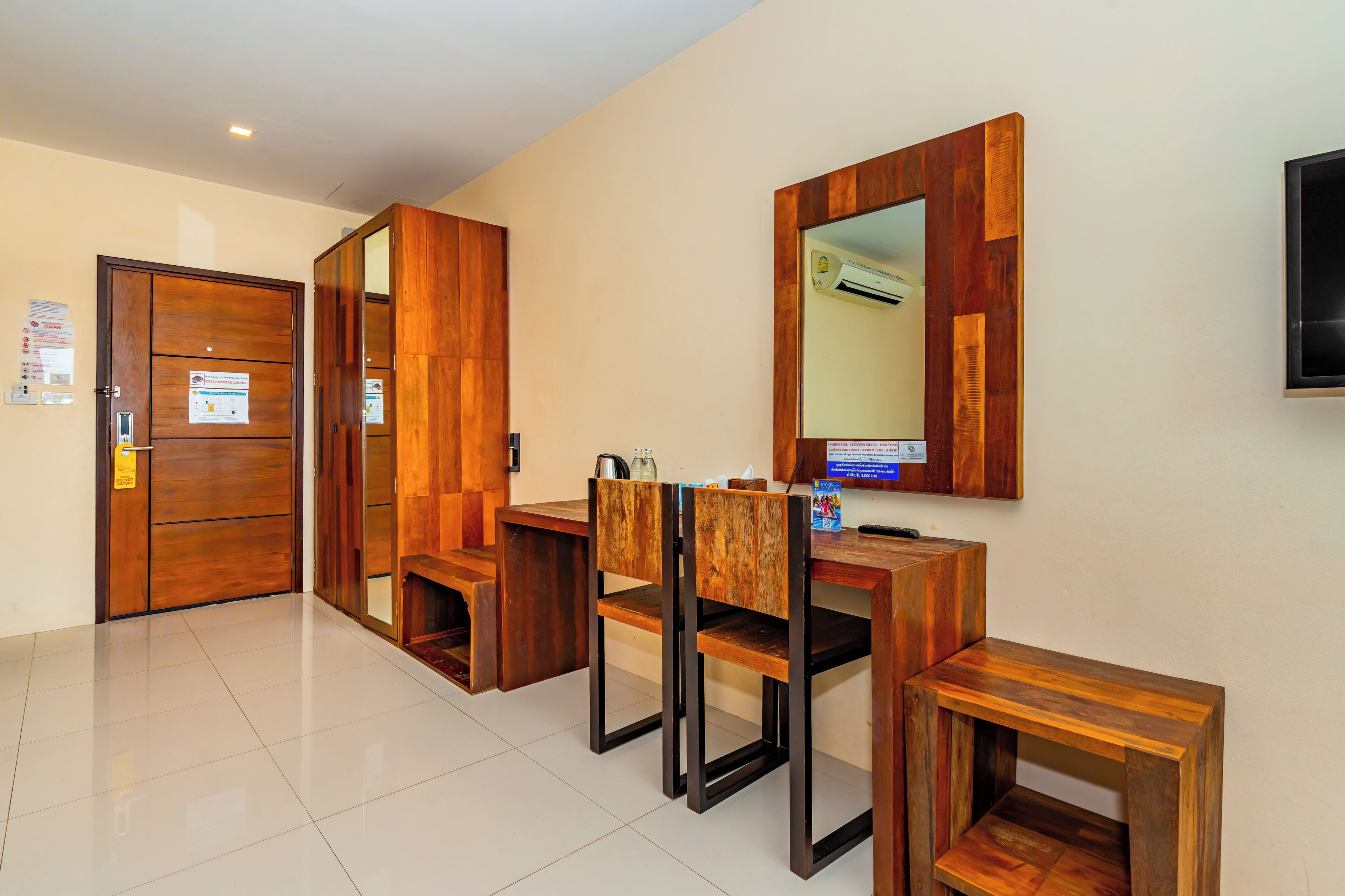 A hotel room with a wooden desk, chairs, a small table, a mirror, and a television on the wall.