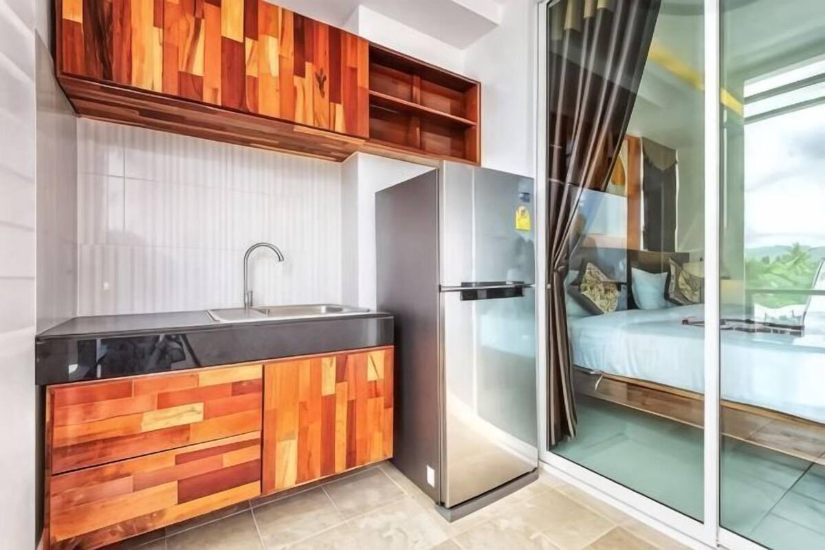 A modern kitchen with a black countertop, wooden cabinets, a stainless steel refrigerator, and a sliding glass door leading to a balcony with a bed.