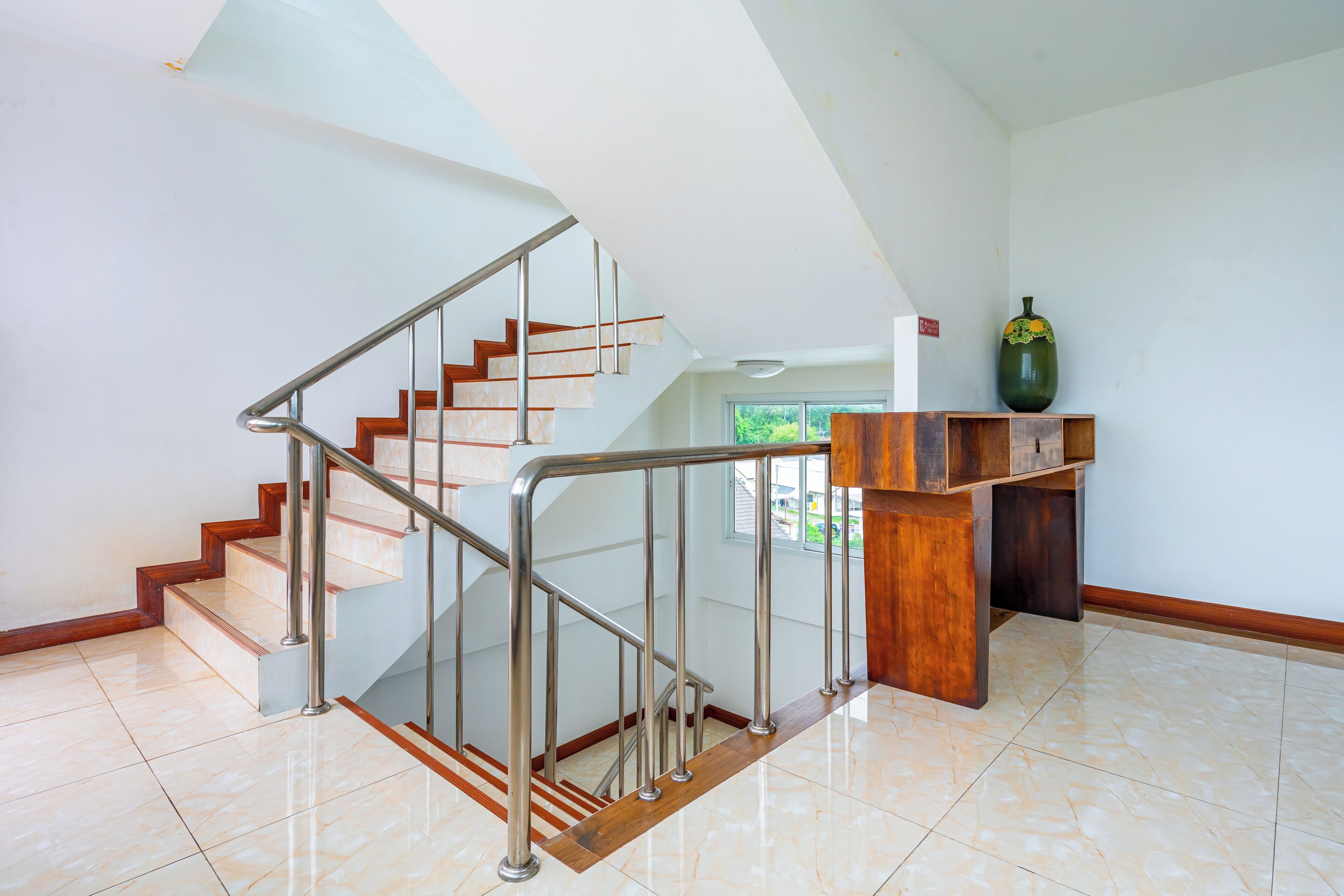 A modern staircase with glass railings and wooden steps, leading to a landing with a wooden podium and a vase.
