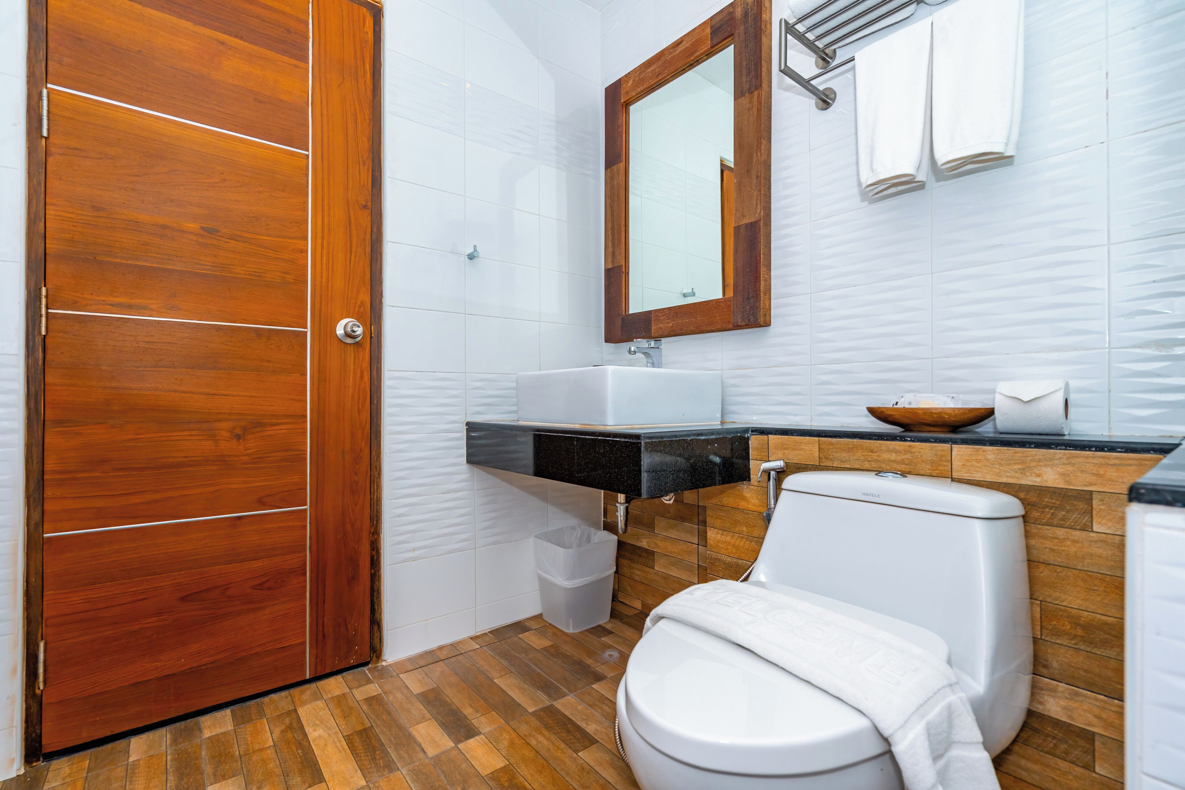 A bathroom with a wooden door, white tiles, a sink, a mirror, and a toilet.