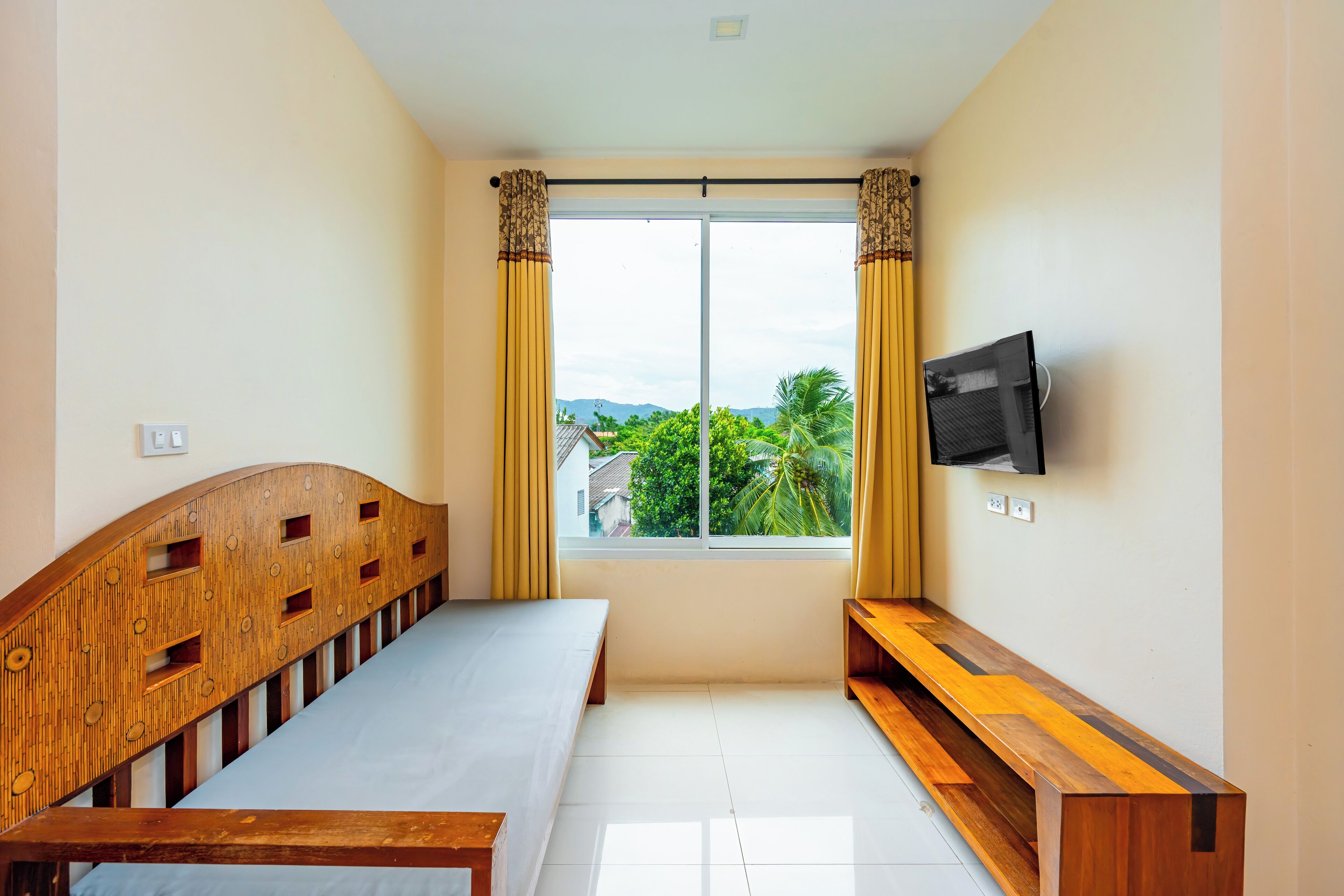 Un dormitorio con cama, un banco, televisor y ventana con cortinas.