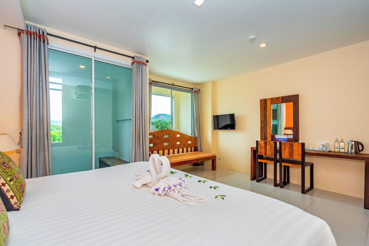 A hotel room with a bed, a wooden bench, a desk with a mirror, and a TV.