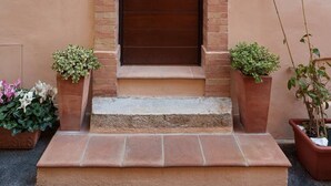 Una puerta de madera marrón con un portón metálico, flanqueada por pilares de ladrillo y un pequeño arco. Hay plantas en macetas a cada lado de la entrada.