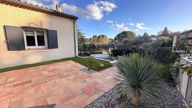 Eine Terrasse mit roten und beigen Fliesen, ein kleiner Garten mit einer Yucca-Pflanze und ein weißes Gebäude mit Fenster und Rollo.