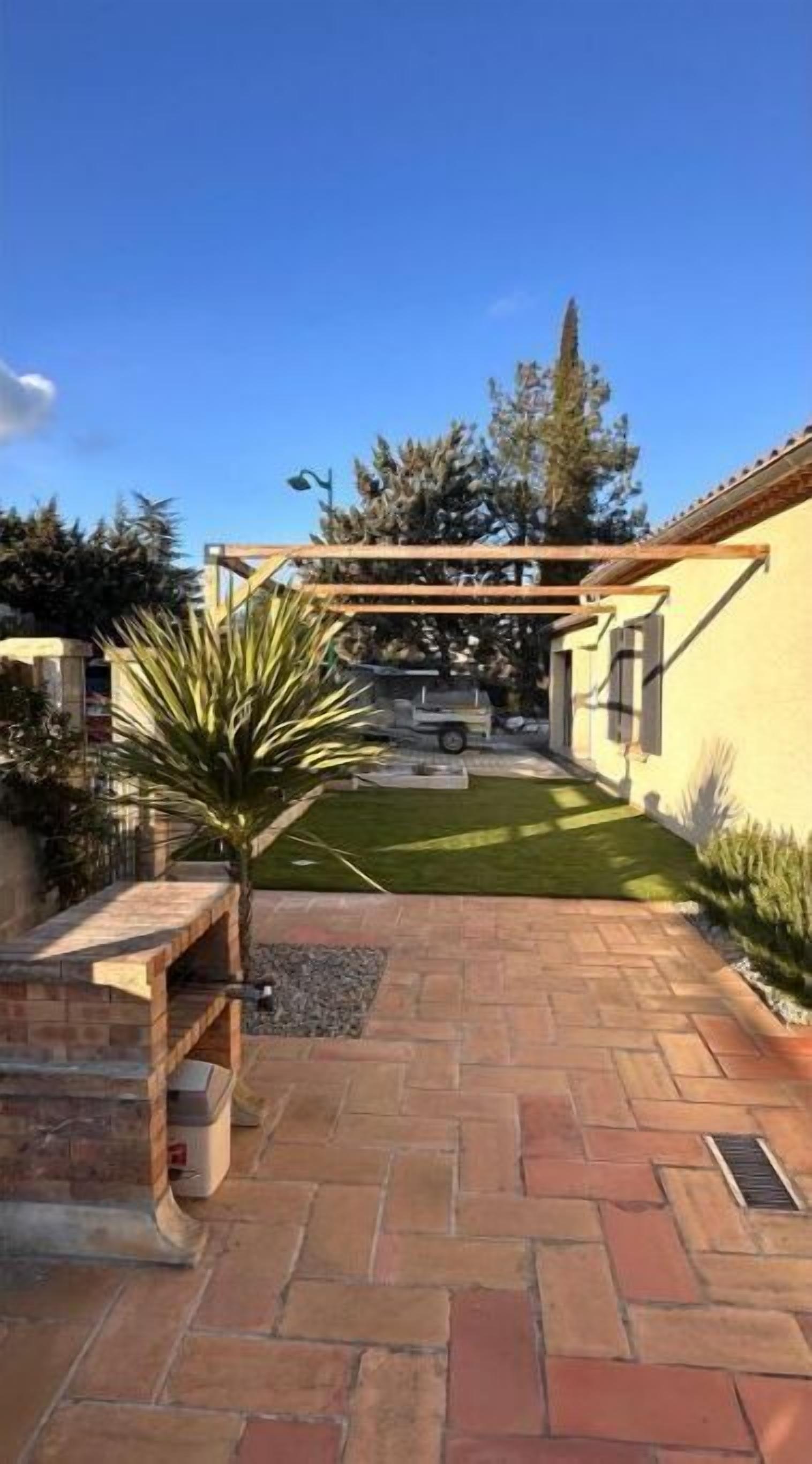 A paved patio area with a pergola structure, a small lawn, and a vehicle in the background.