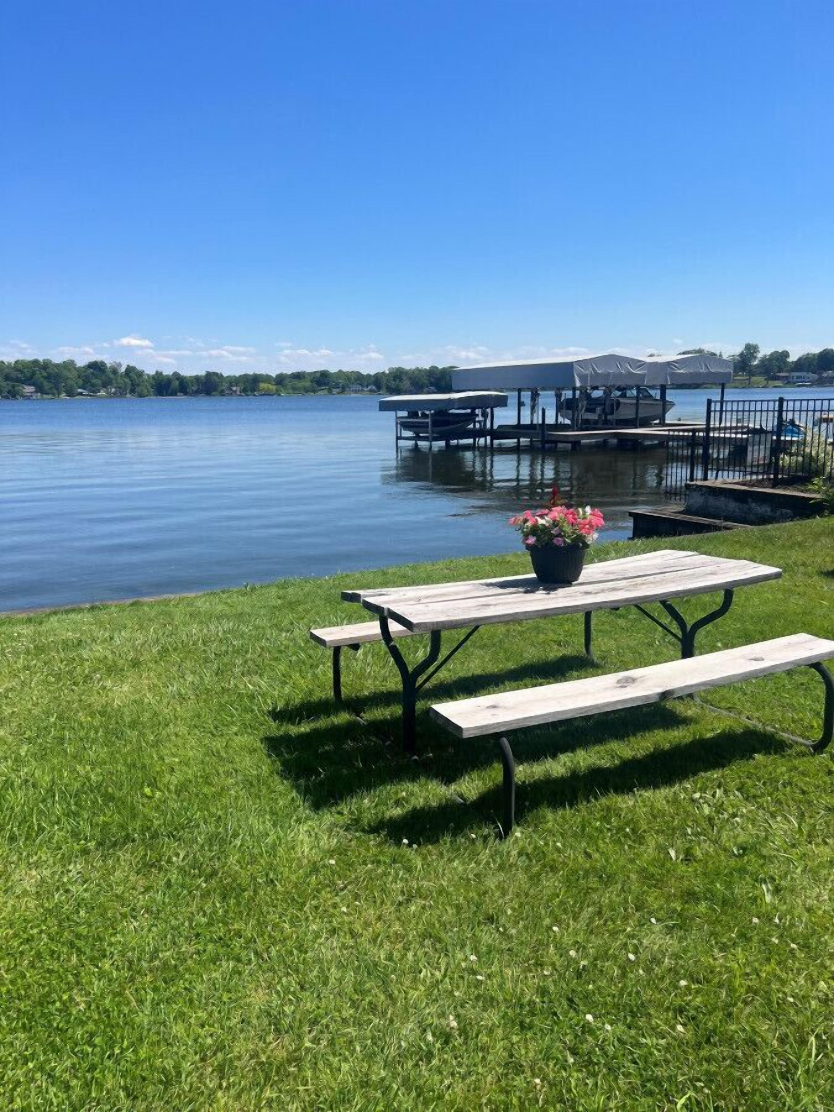 A lakeside picnic area with a wooden bench and picnic table, surrounded by lush green grass.