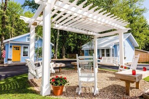 Une pergola blanche avec des sièges, une petite table et un pot de fleurs, située dans un quartier résidentiel aux maisons bleues.