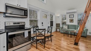 Cocina moderna con armarios blancos, un microondas de acero inoxidable y un horno empotrado. Cuenta con una zona de comedor con sillas de madera y una mesa. El ambiente tiene piso de madera y un sofá verde en la sala.