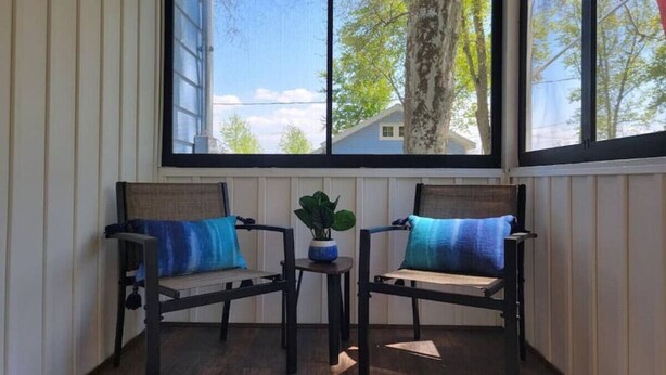 Een kamer met twee houten stoelen met blauwe kussens, een kleine tafel met een plant en een raam met uitzicht op bomen en een huis.