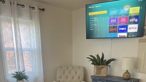 Ett vardagsrum med en soffa, ett soffbord, en fåtölj, ett litet sidobord och en tv som är monterad på väggen och visar gränssnittet för en streamingtjänst.