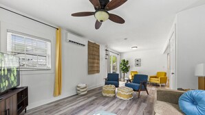 Una sala de estar con televisor, suelo de madera, ventilador de techo y zona de estar con sillones y otomanas.