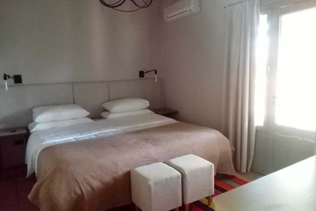 Una habitación de hotel con cama, mesitas de noche, un banco y una ventana con cortinas.
