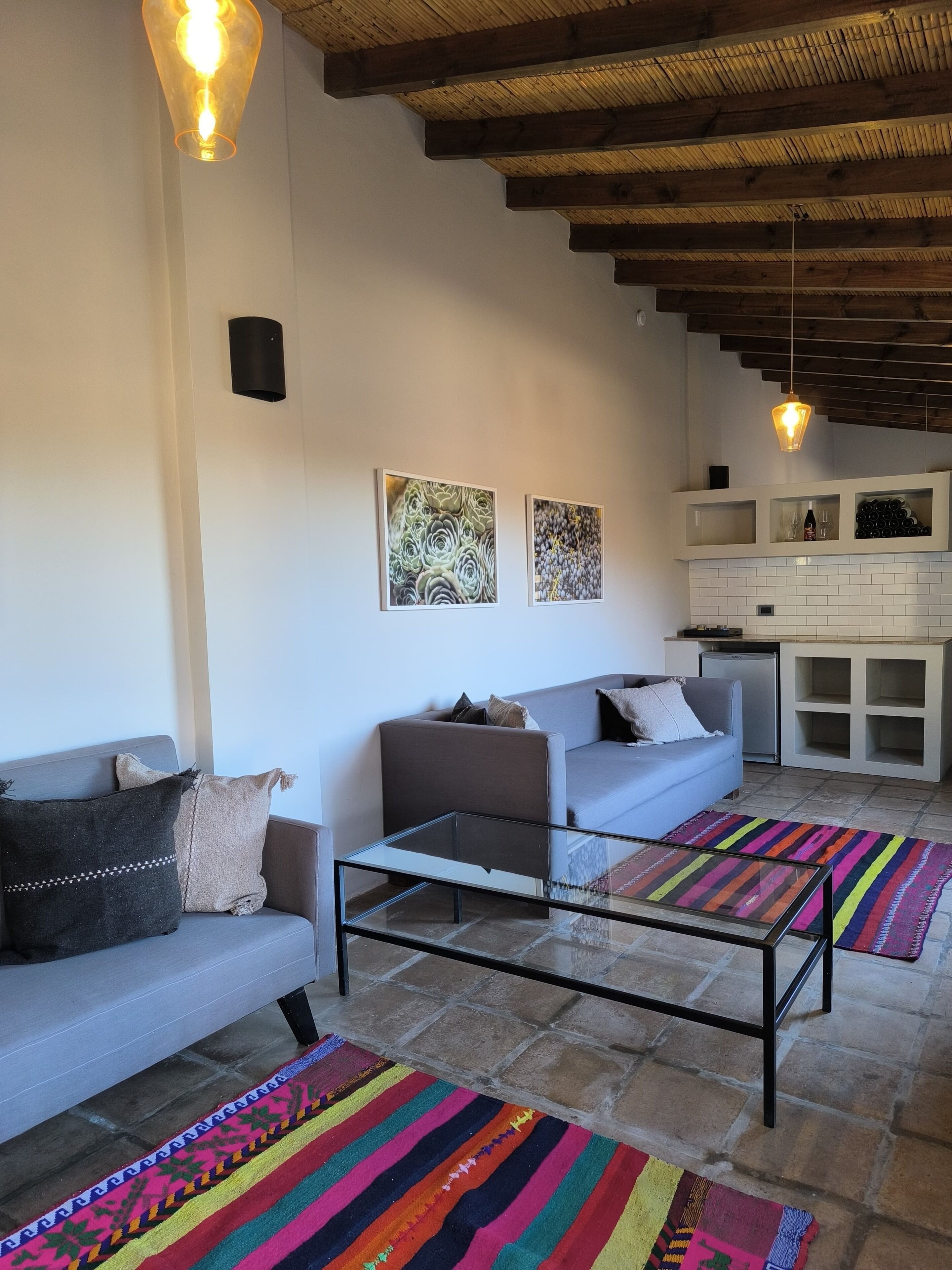A modern living room with a sofa, a glass coffee table, and colorful rugs. There are two wall-mounted artworks and a kitchen area with cabinets and a refrigerator.