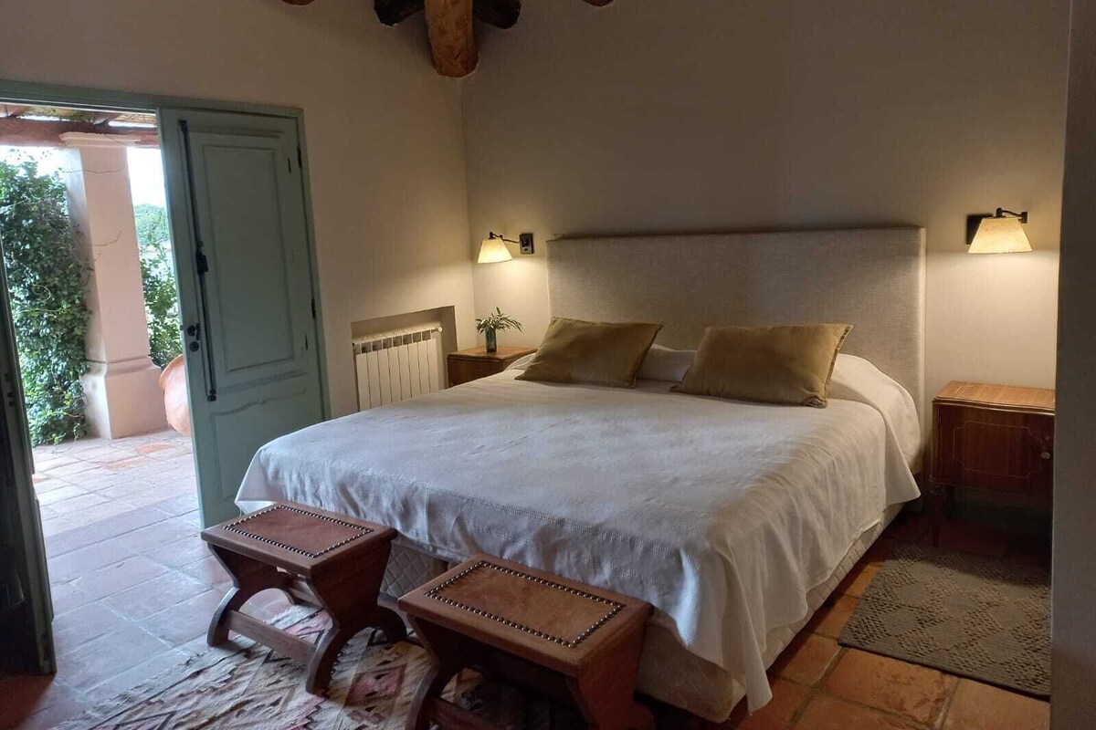 Un dormitorio con una cama grande, dos mesitas de noche, un banco de madera y vistas al exterior.