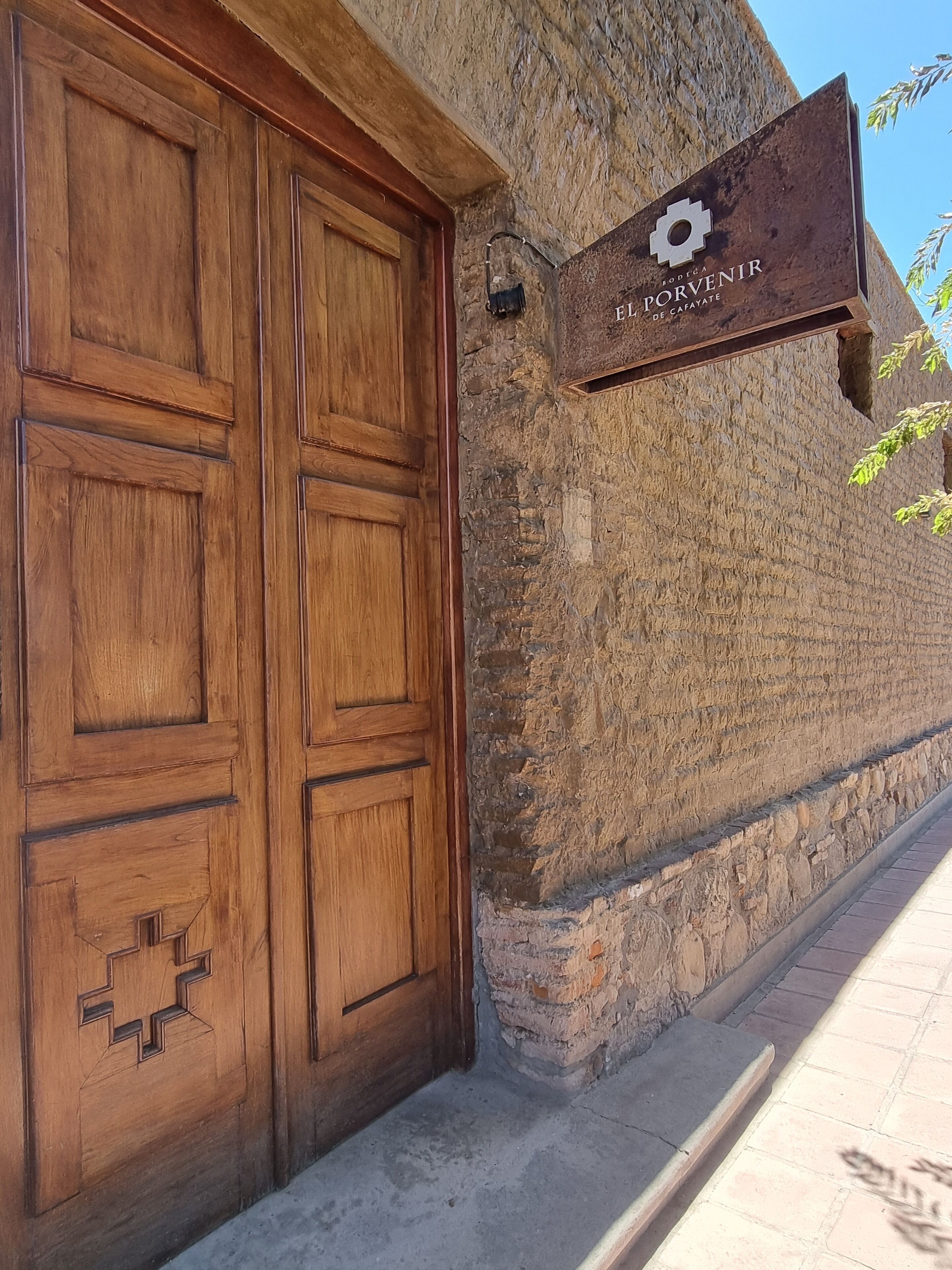 A large wooden door with a geometric design, a stone wall, and a sign with a logo and text.