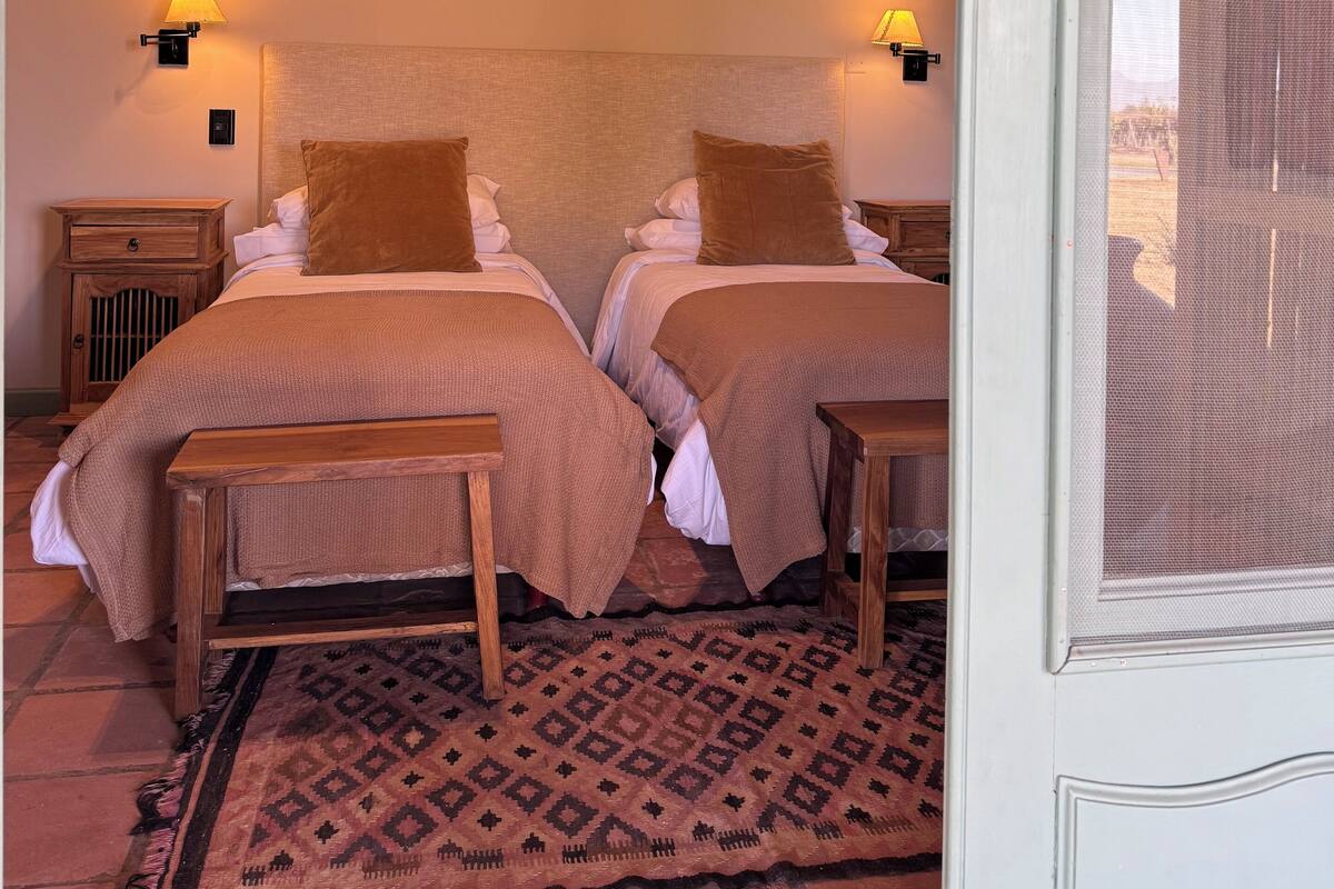 Ein Doppelzimmer mit zwei Betten, Holzkopfteilen, einem gemusterten Teppich und Blick durch ein Fenster mit Moskitonetz.
