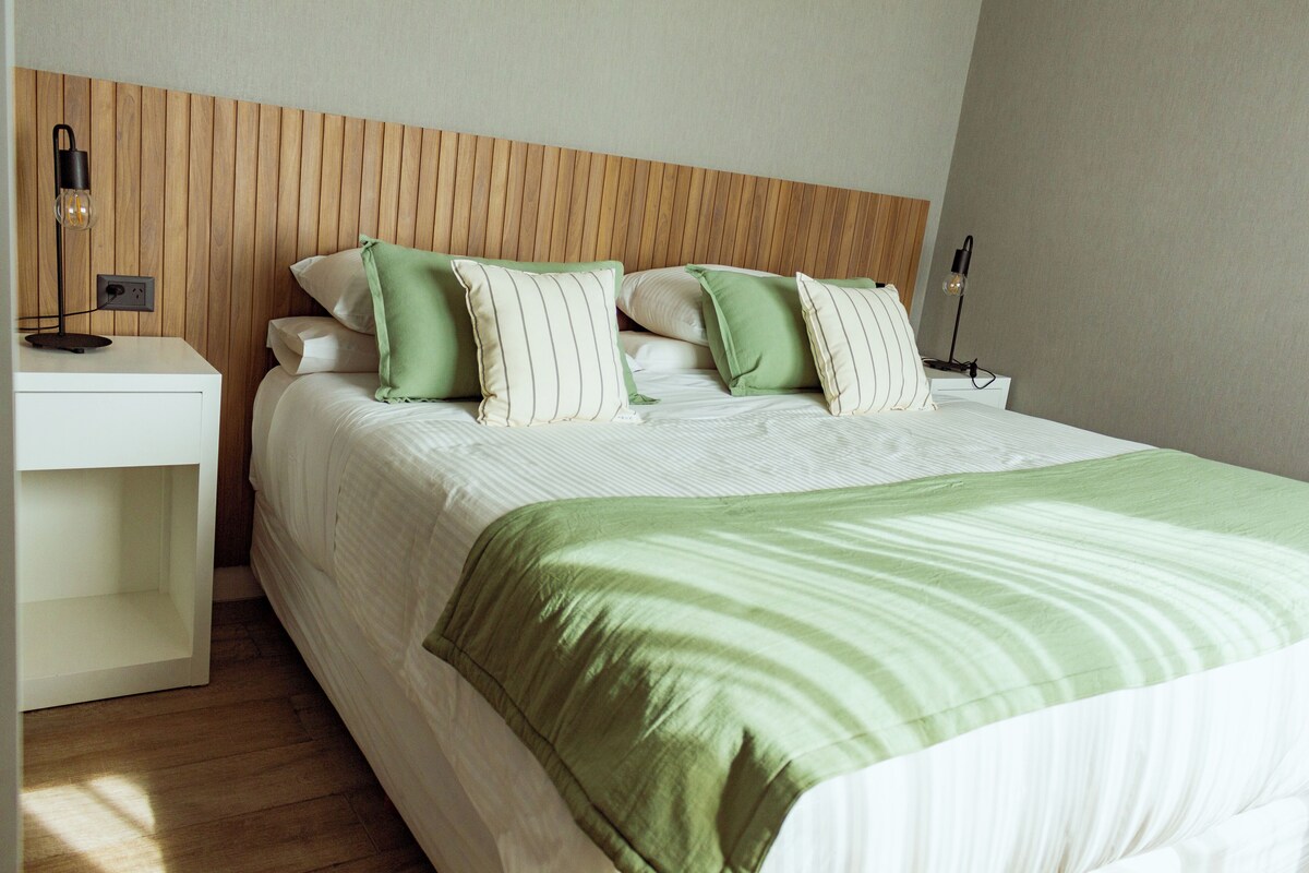 Una cama bien hecha con una colcha verde y blanca a rayas, dos mesitas de noche con lámparas y un cabecero de madera.
