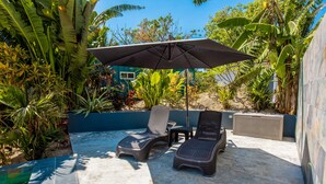 Een terras buiten met twee lounge stoelen en een grote parasol, omgeven door weelderige tropische planten en een zwembad.