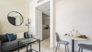 Uma sala de estar moderna com uma mesa de centro de vidro, um sofá com almofadas, um espelho redondo e uma área de jantar com mesa posta para dois.