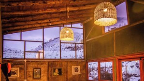 A cozy room with a wooden ceiling, large windows with a snowy view, and a table with books and a patterned tablecloth. - Domos Andinos HostCamp  (Potrerillos)