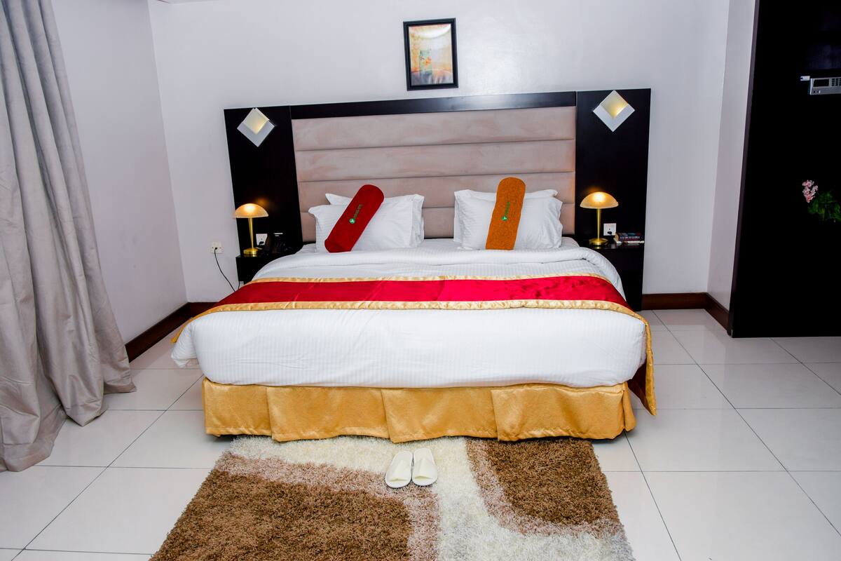 A neatly made bed with a red and orange skateboard, white pillows, and a gold skirted bedspread. There are two bedside lamps, a framed picture on the wall, and a brown shag rug on the floor.