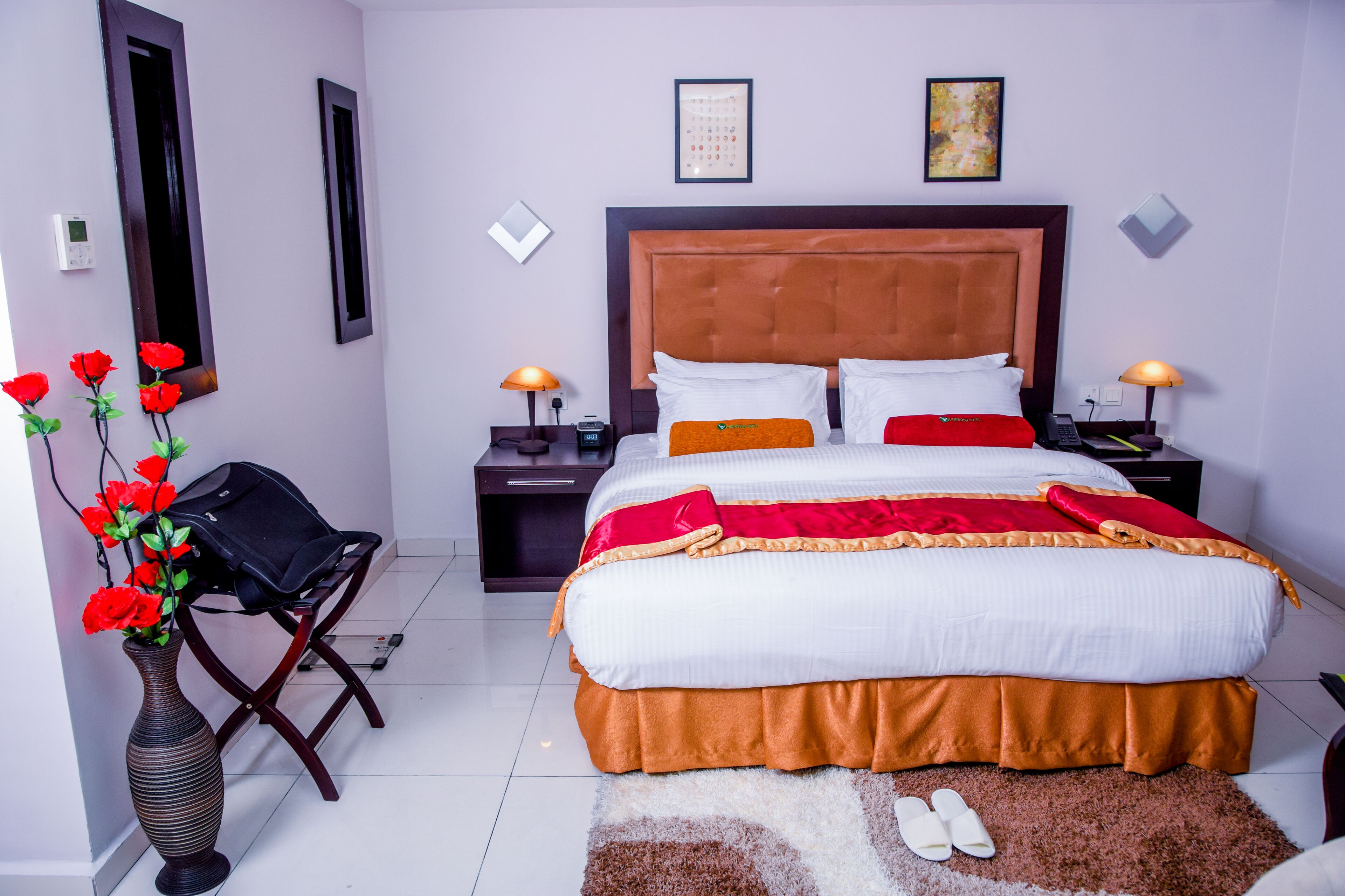 A neatly arranged hotel room with a large bed, two bedside tables, a nightstand, a vase with red flowers, and a pair of shoes on the floor.