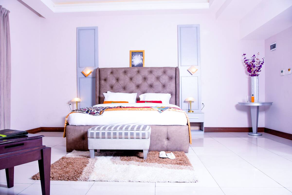 A modern bedroom with a large bed, bedside tables, a nightstand, a small round table, and a vase with flowers.