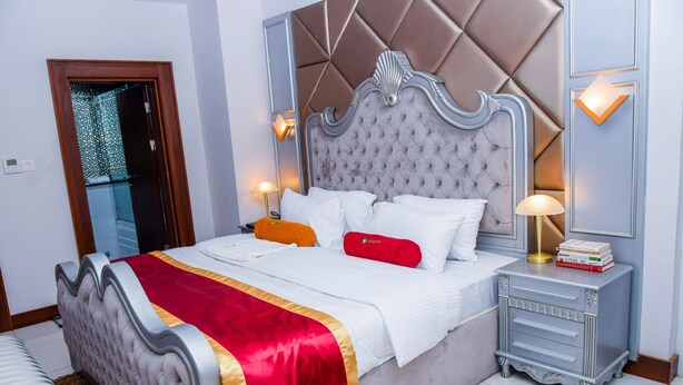 Ein ordentlich bezogenes Bett mit roter und goldener Tagesdecke, einem gepolsterten Kopfteil und Nachttischlampen.
