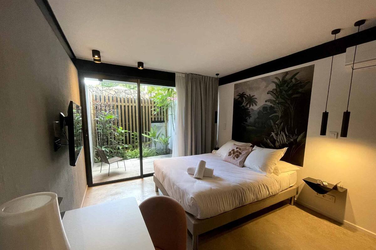 A modern hotel room with a bed, a desk, and a view of a garden through a sliding door.