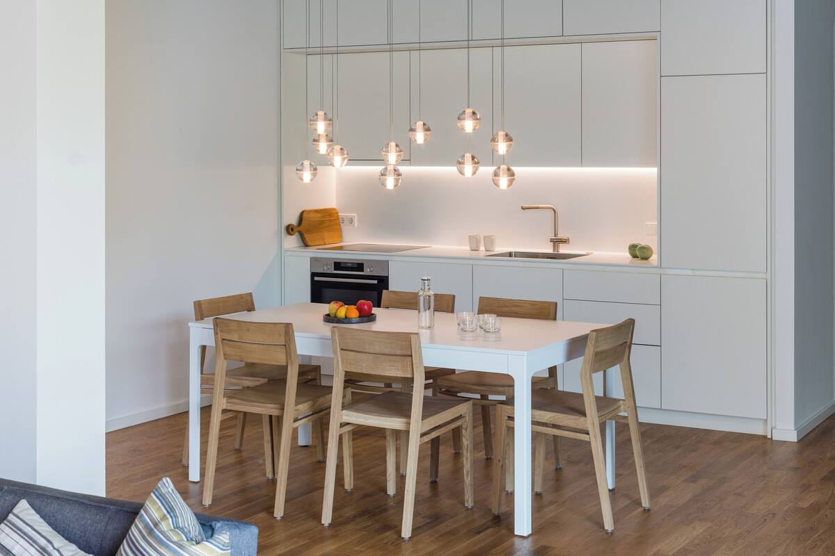 A modern kitchen with a white countertop, wooden chairs, and pendant lights.