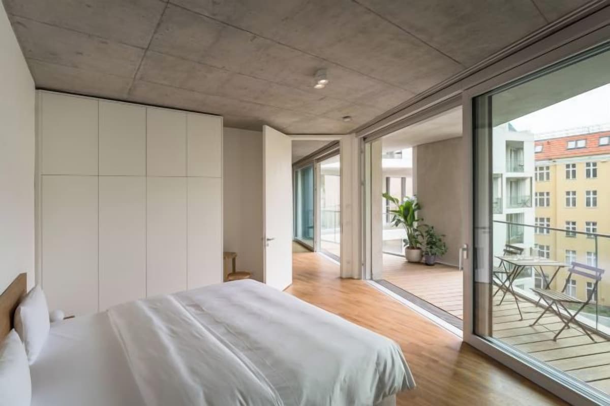 Et soveværelse med en stor seng, en skydedør til en balkon med havemøbler og udsigt over bybilledet.