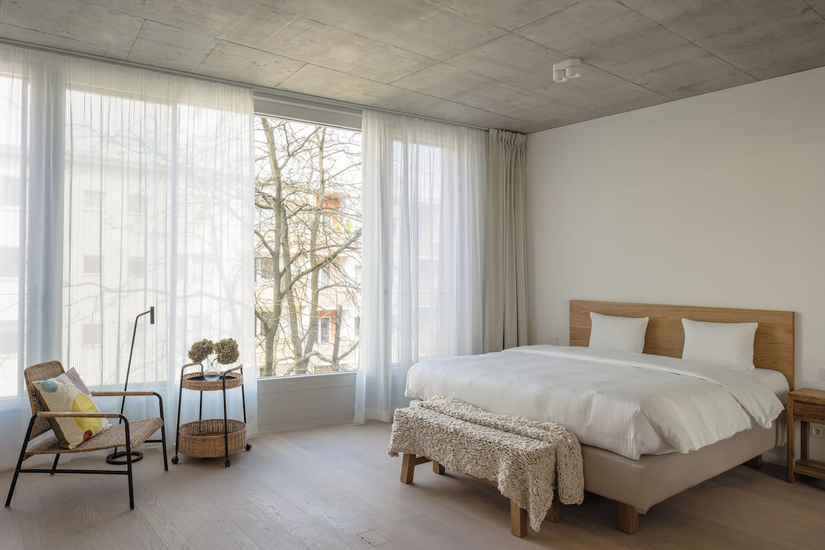 A bedroom with a large bed, a chair, a small table with a plant, and a window with sheer curtains.