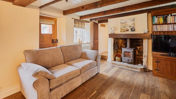 A cozy living room with a beige sofa, a stone fireplace, wooden flooring, and a bookshelf. - Host & Stay | Colcott Cottage (Fylingthorpe)