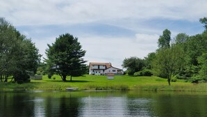 A house with a pond in front, surrounded by trees and grass. - Lakefront Escape -1 acre / private pool / kayak / fire pit / swing set (Blakeslee)