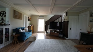 A spacious living room with a blue sofa, a flat-screen TV, and a wooden cabinet. - Lakefront Escape -1 acre / private pool / kayak / fire pit / swing set (Blakeslee)