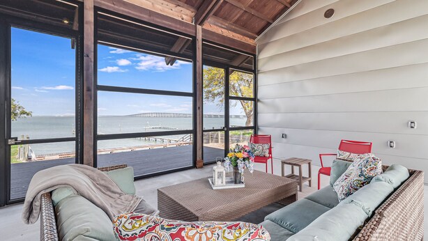 A spacious room with large windows overlooking a body of water, a wicker sofa, a red chair, and a small table with a vase of flowers.