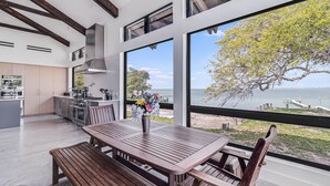 A modern kitchen with a large island, a dining area with a wooden table and bench, and a view of the sea through large windows.