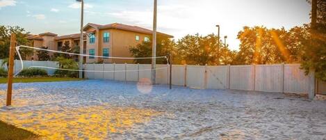 Un terrain de volleyball de sable avec un filet, entouré d’une petite clôture et avec un bâtiment en arrière-plan.