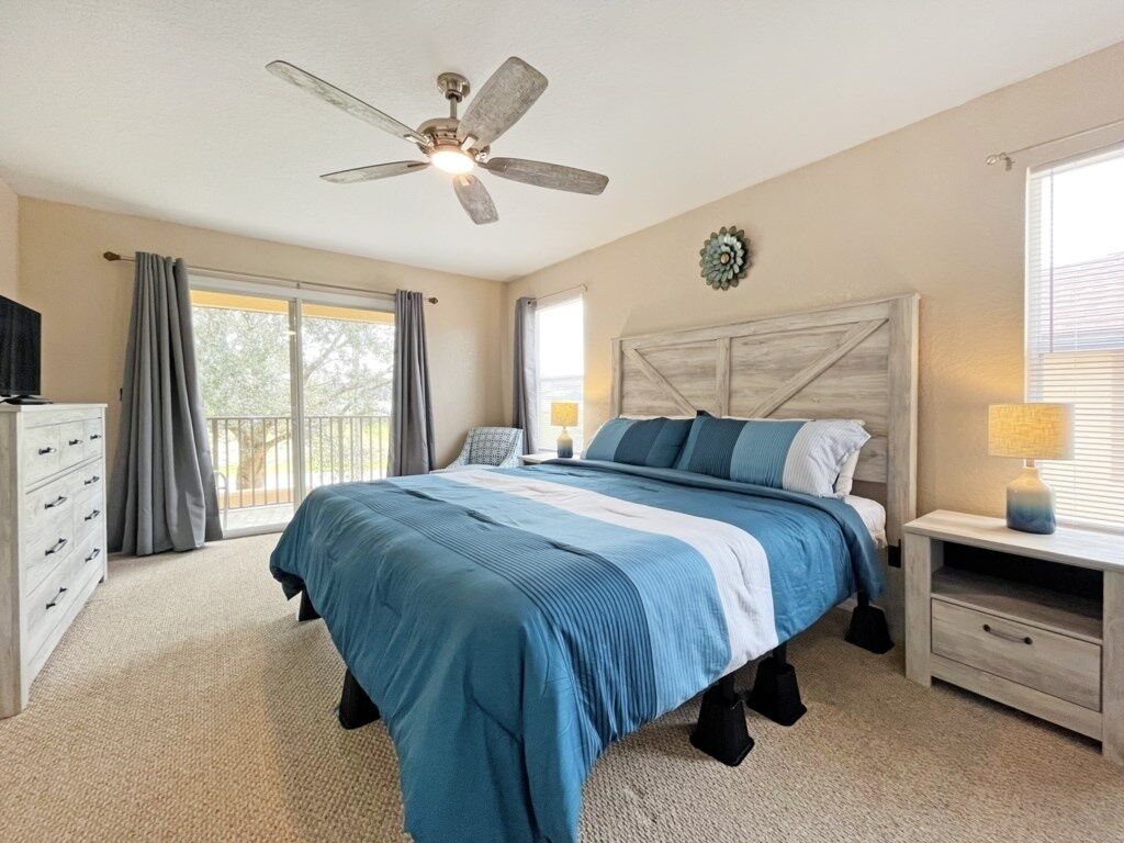 Um quarto com cama, mesa de cabeceira, cômoda, ventilador de teto e janela com cortinas.