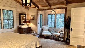 A cozy bedroom with a large bed, a dresser, two chairs, and a window with a view of trees. - Luxury Lake Rabun Cabin - restored lakefront w/ stunning views fr/every room! (Lakemont)
