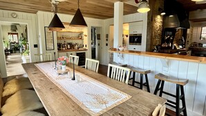 A wooden dining table with a lace runner, white chairs, and a bar area with stools. - Luxury Lake Rabun Cabin - restored lakefront w/ stunning views fr/every room! (Lakemont)