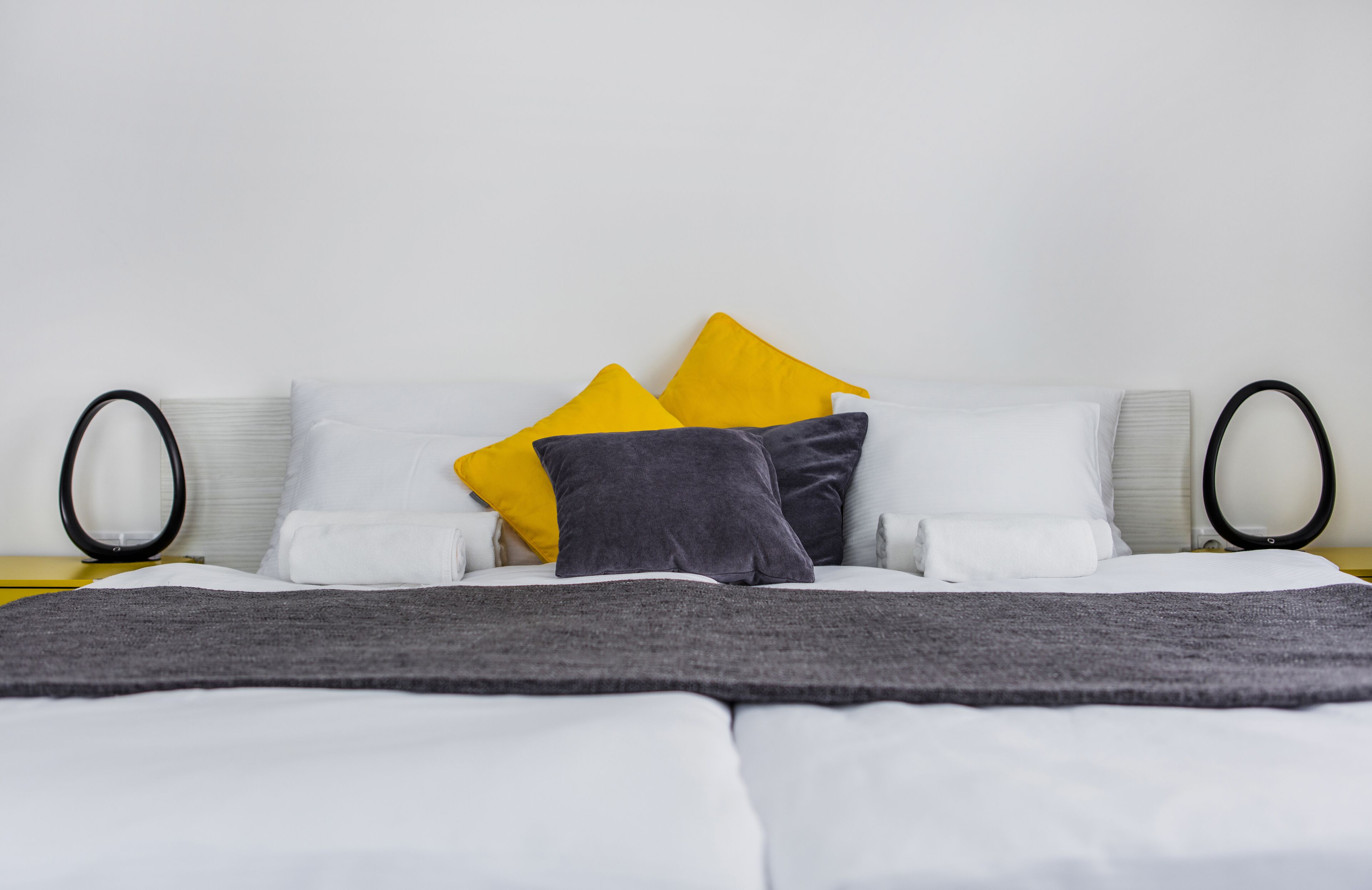 Ein ordentlich gemachtes Bett mit weißer und grauer Bettwäsche, zwei gelbe Kissen und einer grauen Tagesdecke.