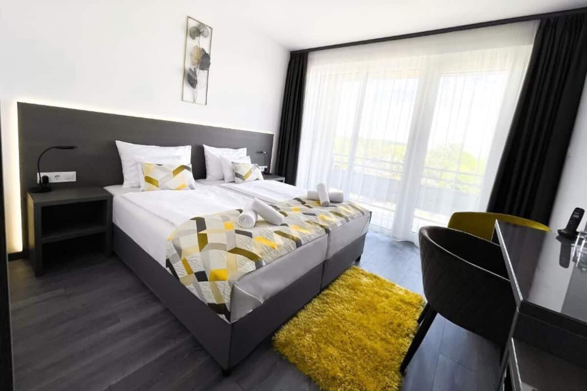 A modern hotel room with a large bed, a nightstand, a chair, and a view of the outdoors through large windows.