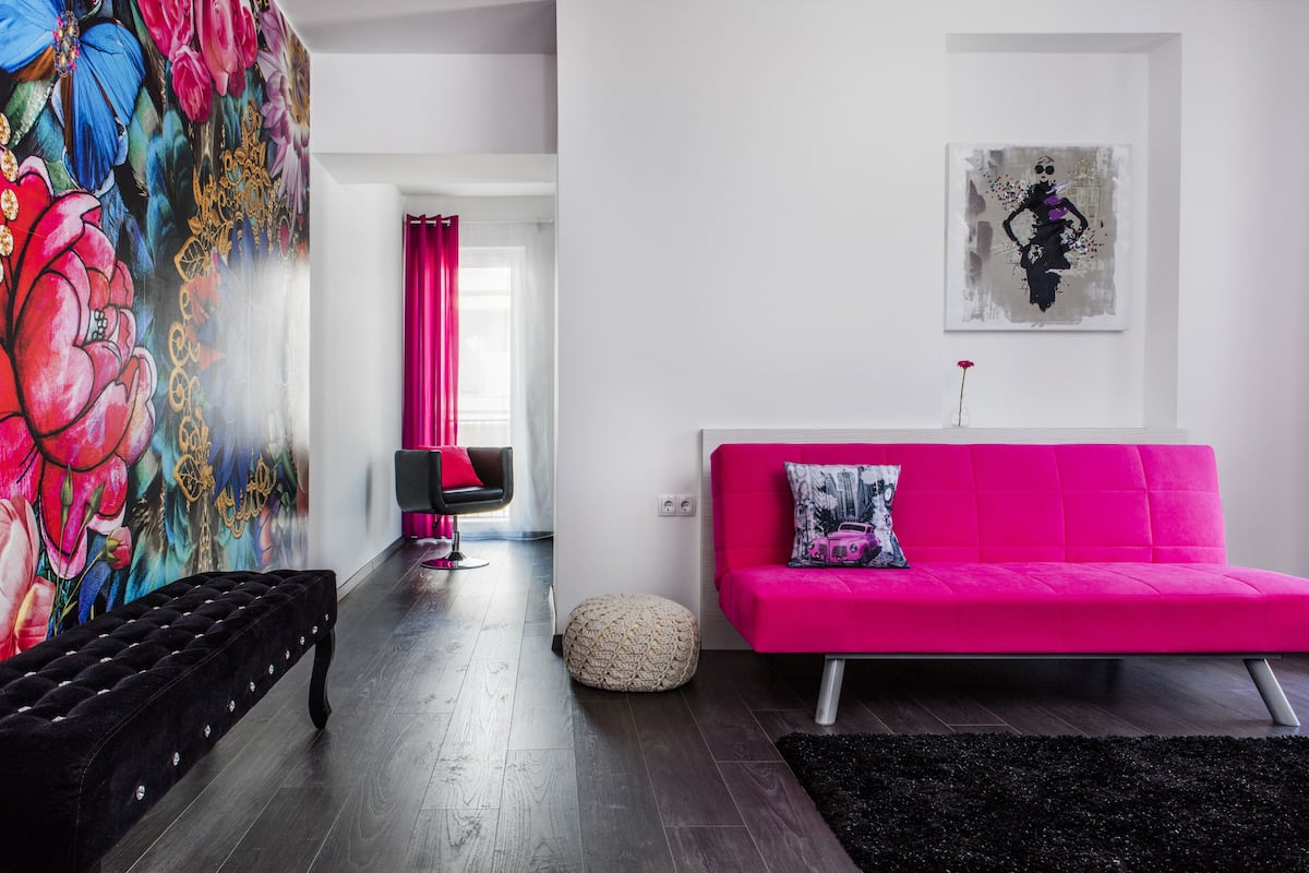 A modern living room with a vibrant pink sofa, a tufted black bench, and a colorful floral mural on the wall.
