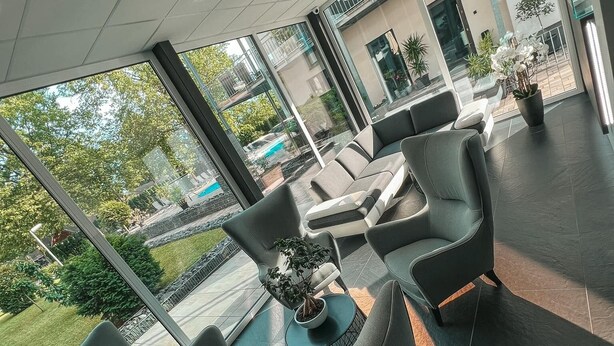 A modern indoor patio area with a seating arrangement, large windows, and a view of a swimming pool and greenery outside. - Hotel Csopak Resort & Lake (Csopak)