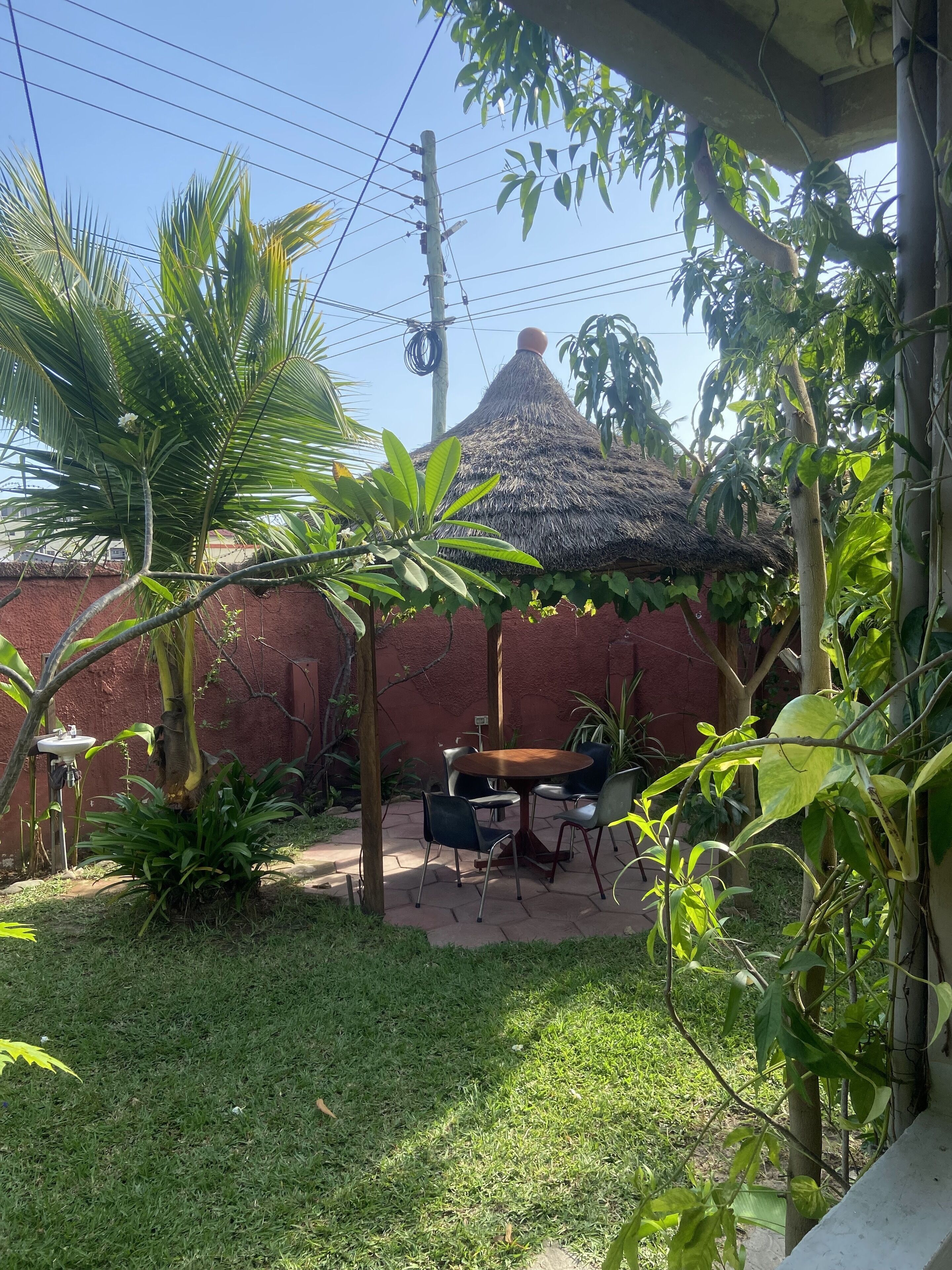 A patio area with a thatched-roof structure, surrounded by lush greenery and a red wall.