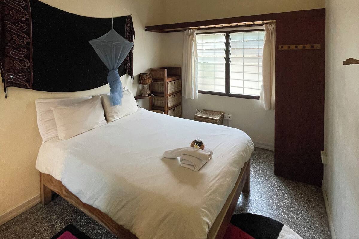 Una habitación de hotel con cama, ventana con cortinas, mesita de noche con lámpara y un pequeño armario de madera.