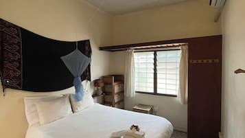 Una habitación de hotel con cama, ventana con cortinas, mesita de noche con lámpara y un pequeño armario de madera.
