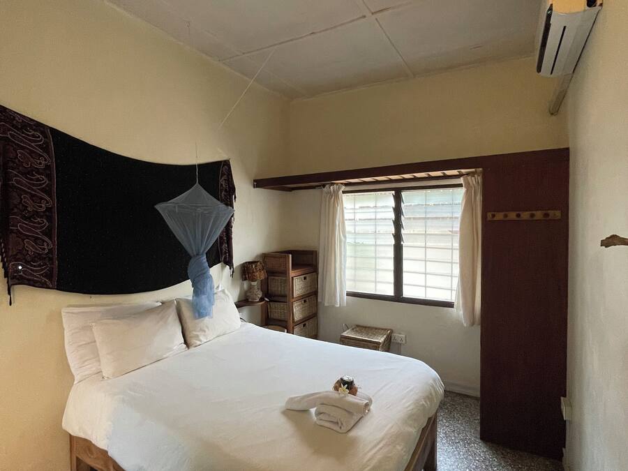 Una habitación de hotel con cama, ventana con cortinas, mesita de noche con lámpara y un pequeño armario de madera.